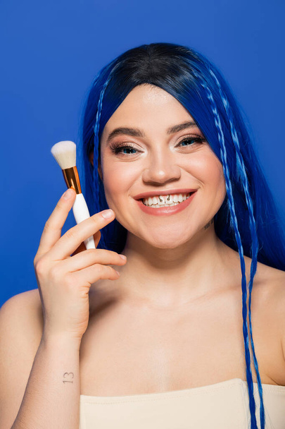 individualism, joyous young woman with vibrant eyes and hair looking at camera while holding makeup brush on blue background, cosmetic, self expression, visage, youth, beauty trends  - Foto, Imagem