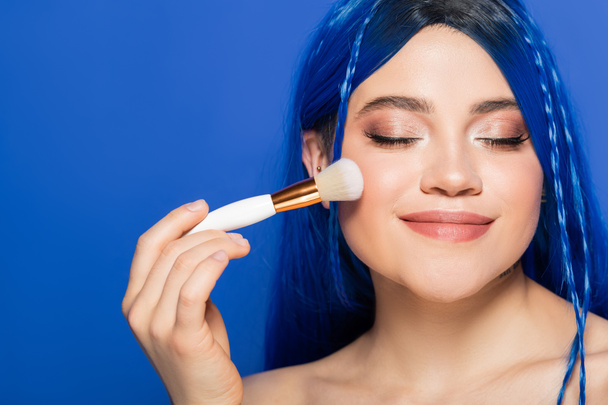 individualism, pleased young woman with vibrant eyes and hair looking at camera while holding cosmetic brush on blue background, applying makeup, self expression, visage, youth, beauty trends  - Photo, Image