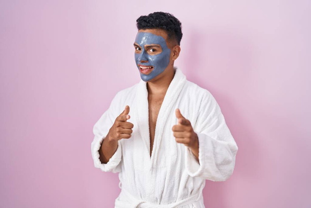 Молодий іспаномовний чоловік носить маску для обличчя краси та халат для ванни, вказуючи пальці на камеру з щасливим та смішним обличчям. хороша енергія і вібрує.  - Фото, зображення