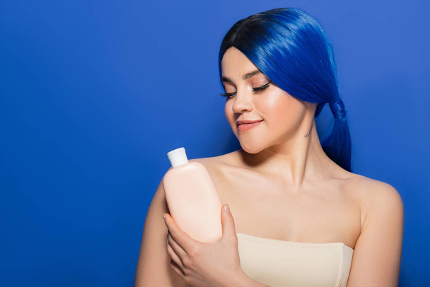 concept de soins du corps et des cheveux, portrait de jeune femme tatouée avec une couleur de cheveux vibrante posant avec des épaules nues sur fond bleu, tenant bouteille cosmétique avec shampooing, tendances beauté  - Photo, image