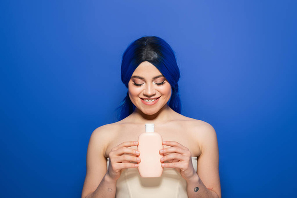 concept de soins capillaires, portrait d'une jeune femme gaie avec une couleur de cheveux vibrante posant avec des épaules nues sur fond bleu, tenant bouteille cosmétique avec shampooing, tendances de la beauté, cheveux sains  - Photo, image