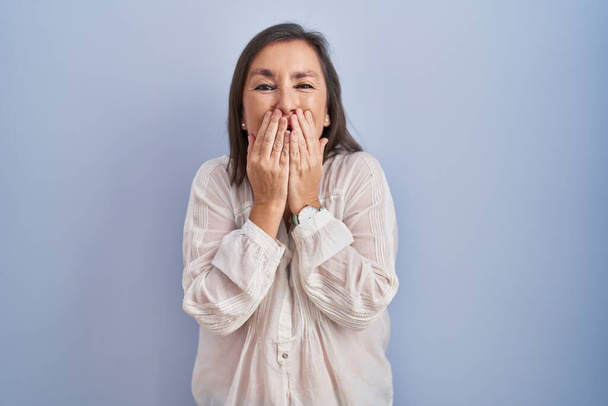 Middle age hispanic woman standing over blue background laughing and embarrassed giggle covering mouth with hands, gossip and scandal concept  - Фото, зображення