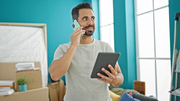 Young hispanic man talking on smartphone using touchpad at new home - Photo, Image