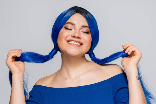 positivity and youth, tattooed woman with closed eyes and dyed hair smiling on grey background, hairstyle, blue hair, modern beauty, self expression, individualism  - Valokuva, kuva