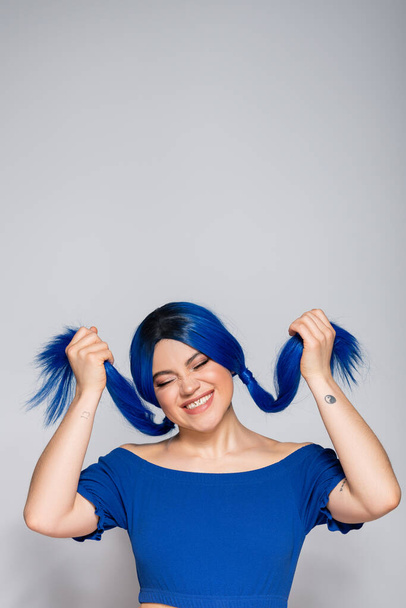 positivity and youthful energy, tattooed and happy woman with closed eyes and dyed hair smiling on grey background, hairstyle, blue hair, modern beauty, self expression, individualism  - Foto, Bild