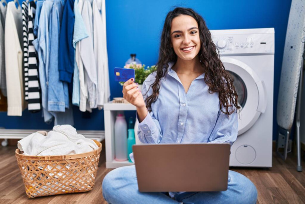 Young hispanic woman using laptop and credit card waiting for washing machine at laundry room - Фото, зображення