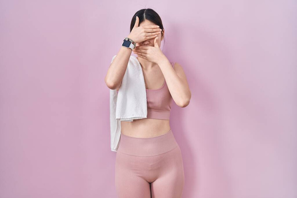 Chinese young woman wearing sportswear and towel covering eyes and mouth with hands, surprised and shocked. hiding emotion  - Fotó, kép
