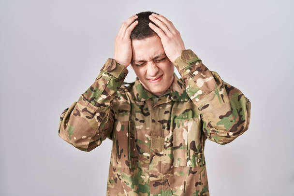 Kamuflaj üniforması giyen genç adam acı ve migren yüzünden çaresiz ve stresli bir şekilde baş ağrısı çekiyor. Eller başının üstünde..  - Fotoğraf, Görsel