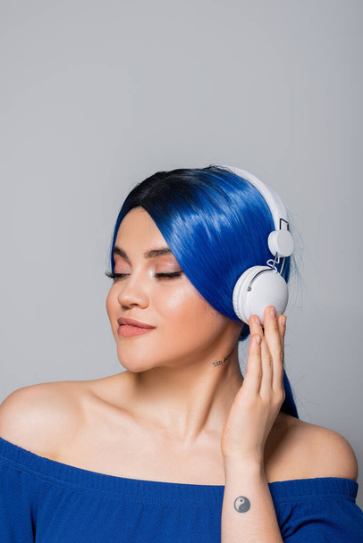zelfexpressie, muziekliefhebber, jonge vrouw met blauw haar luistermuziek in draadloze koptelefoon op grijze achtergrond, gesloten ogen, levendige jeugd, individualisme, moderne subcultuur, tatoeage, geluid  - Foto, afbeelding