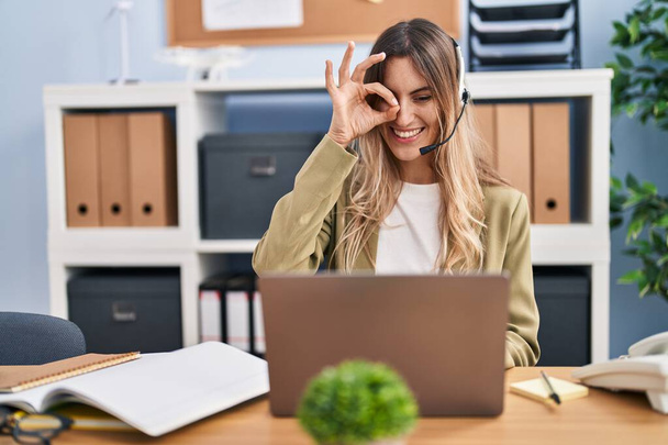 Mujer hispana joven usando auriculares de agente de centro de llamadas trabajando en la oficina sonriendo feliz haciendo signo de ok con la mano en el ojo mirando a través de los dedos  - Foto, imagen