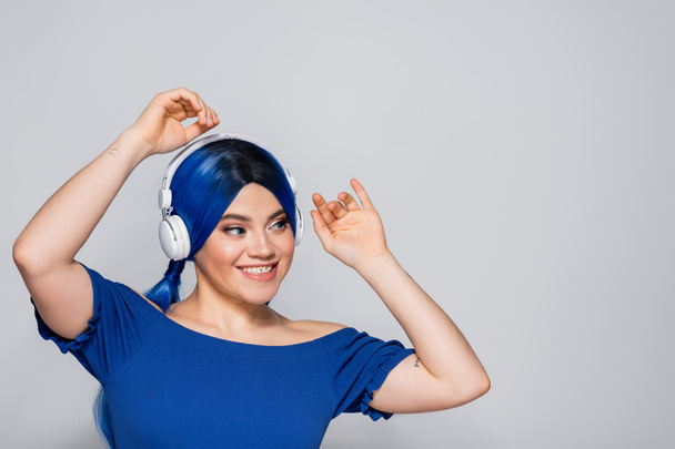 Selbstausdruck, positive junge Frau mit blauen Haaren, die Musik in drahtlosen Kopfhörern auf grauem Hintergrund hört, lebendige Jugend, Individualismus, moderne Subkultur, Tätowierung, Sound  - Foto, Bild