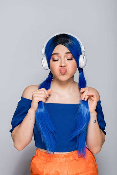 самовираження, молода жінка з блакитним волоссям слухає музику в бездротових навушниках на сірому фоні, гладить губи, яскрава молодь, індивідуалізм, сучасна субкультура, татуювання, звук  - Фото, зображення