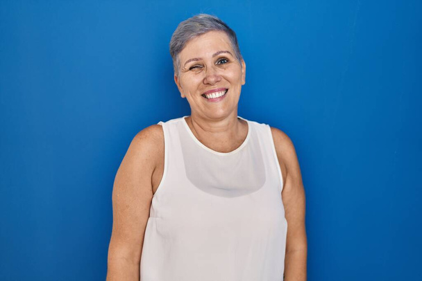 Orta yaşlı beyaz bir kadın mavi arka planda durmuş kameraya seksi bir ifadeyle bakıyor, neşeli ve mutlu bir yüzle..  - Fotoğraf, Görsel