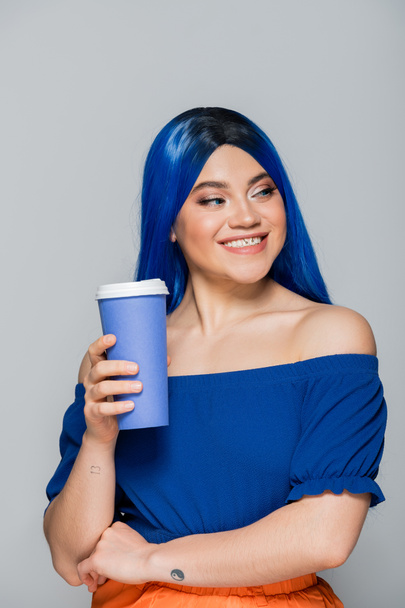 positivity, disposable cup, young woman with blue hair and eyes holding coffee to go on grey background, takeaway, caffeine, energy, tattoo, vibrant color, self expression, individualism  - 写真・画像
