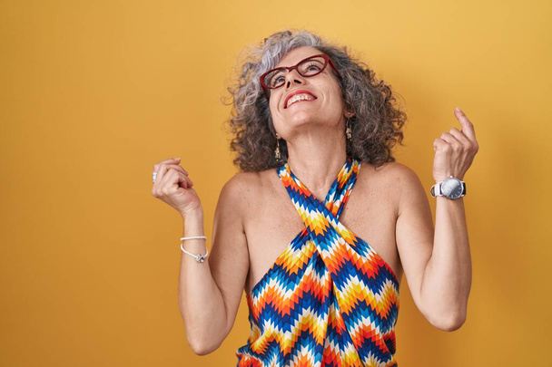Middle age woman with grey hair standing over yellow background celebrating surprised and amazed for success with arms raised and eyes closed. winner concept.  - Fotografie, Obrázek