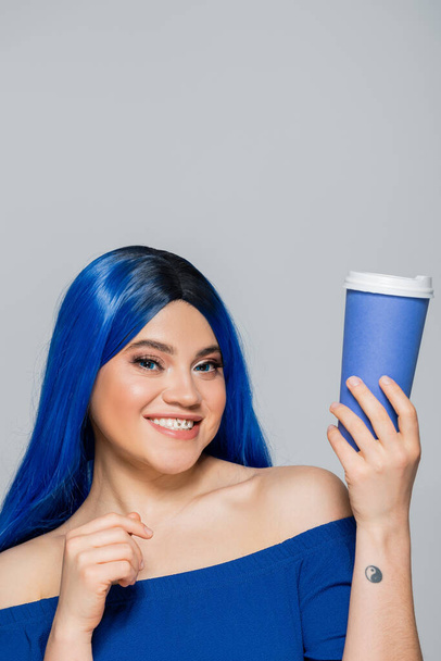 paper cup, happy young woman with blue hair and eyes holding coffee to go on grey background, takeaway, caffeine, energy, tattoo, vibrant color, self expression, individualism  - Foto, immagini