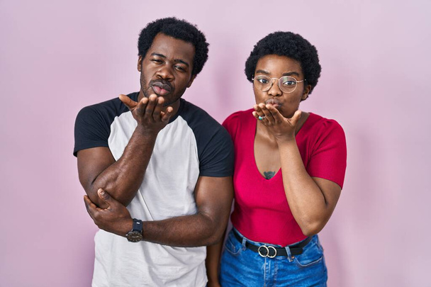 Ein junges afrikanisch-amerikanisches Paar, das vor rosa Hintergrund steht und in die Kamera blickt, bläst einen Kuss mit der Hand in die Luft, der schön und sexy ist. Liebesausdruck.  - Foto, Bild