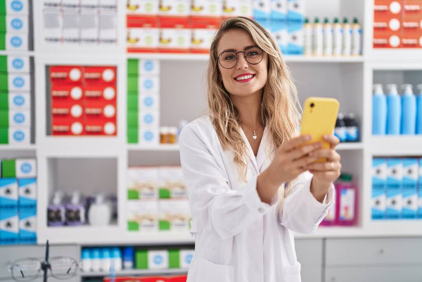 Νεαρή γυναίκα φαρμακοποιός χαμογελά αυτοπεποίθηση χρησιμοποιώντας smartphone στο φαρμακείο - Φωτογραφία, εικόνα