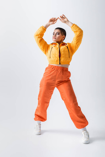 outerwear, casual attire, puffer jacket and trendy orange pants, young tattooed model posing with raised hands on grey background, woman with short hair, generation z - Фото, изображение