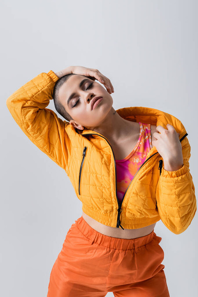 outerwear, individualism, fashion model posing with closed eyes, young woman with short hair standing in yellow puffer jacket on grey background, isolated, youth culture, casual wear - Foto, Bild