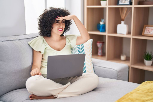 Νεαρή μελαχρινή γυναίκα με σγουρά μαλλιά χρησιμοποιώντας φορητό υπολογιστή κάθεται στον καναπέ στο σπίτι πολύ χαρούμενος και χαμογελώντας κοιτάζοντας μακριά με το χέρι πάνω από το κεφάλι. Αναζήτηση έννοιας.  - Φωτογραφία, εικόνα