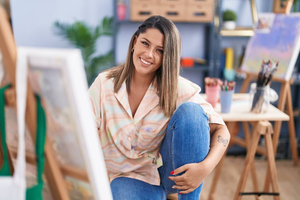 Joven artista hispana sonriendo confiada sentada en el piso del estudio de arte - Foto, imagen