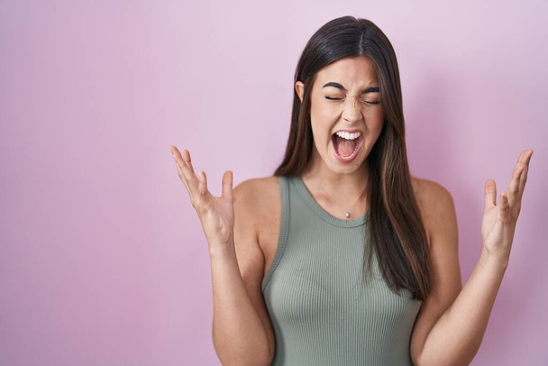 Ισπανίδα γυναίκα στέκεται πάνω από ροζ φόντο γιορτάζει τρελός και τρελός για την επιτυχία με τα χέρια ψηλά και κλειστά μάτια ουρλιάζοντας ενθουσιασμένος. έννοια νικητή  - Φωτογραφία, εικόνα