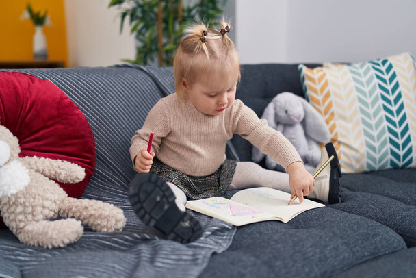Adorable blonde girl preschool student sitting on sofa drawing on notebook at home - Photo, Image