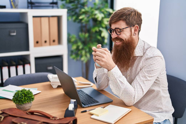 Νεαρή κοκκινομάλλα επιχειρηματίας χρησιμοποιώντας φορητό υπολογιστή πίνοντας καφέ στο γραφείο - Φωτογραφία, εικόνα