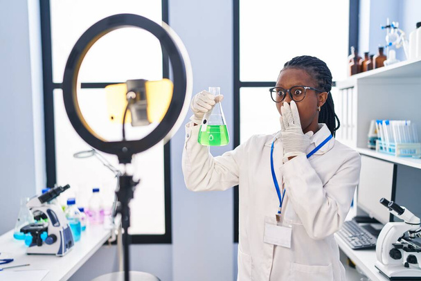 Afrikanerin mit Zöpfen, die im Labor arbeitet und mit dem Smartphone den Mund mit der Hand bedeckt, schockiert und ängstlich vor Fehlern. Überraschte Miene  - Foto, Bild