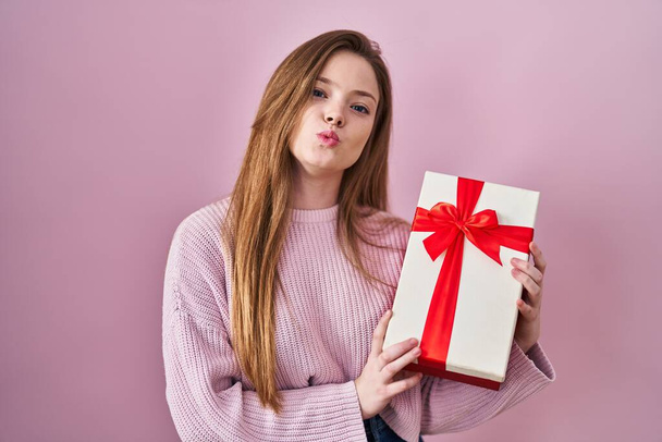 Jonge blanke vrouw die een cadeau vasthoudt, kijkend naar de camera die een kus geeft, omdat ze lief en sexy is. liefde uitdrukking.  - Foto, afbeelding