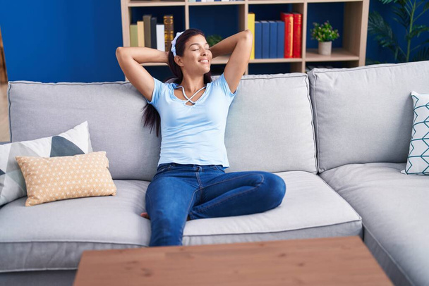 Молода красива іспаномовна жінка розслаблена руками на голові, сидячи на дивані вдома
 - Фото, зображення