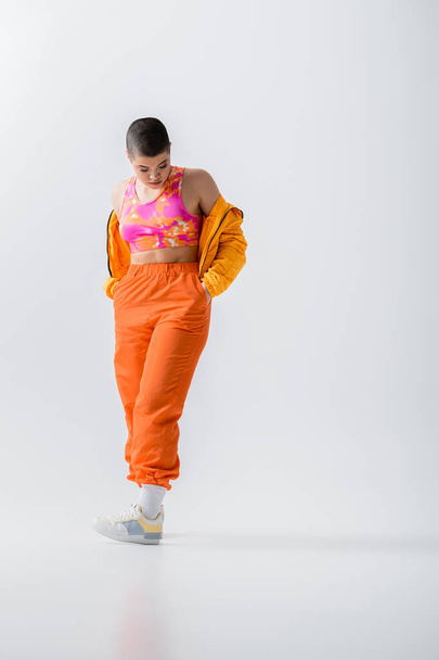 modern subculture, fashionable young woman with short pair posing with hands in pockets of orange pants on grey background, puffer jacket. outerwear, youthful energy, full length  - 写真・画像