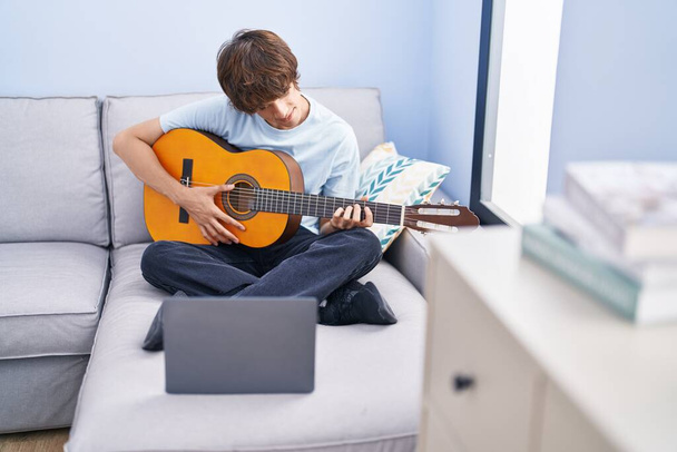 Young blond man having online classical guitar class sitting on sofa at home - Fotó, kép
