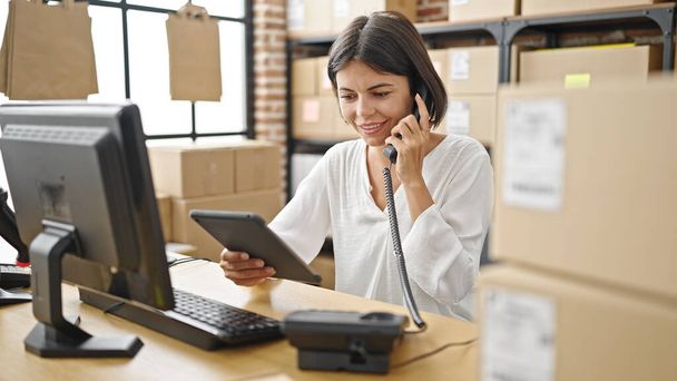 Young beautiful hispanic woman ecommerce business worker talking on telephone using touchpad at office - Foto, Imagen