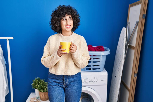 Young middle east woman drinking coffee waiting for washing machine at laundry - Photo, image