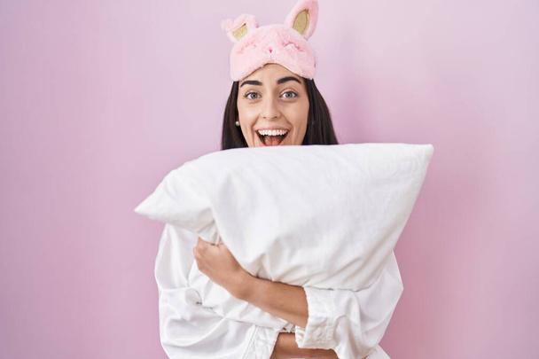 Молодая брюнетка в маске для сна и пижаме, обнимающая подушку, празднующая безумие и пораженная успехом с открытыми глазами, кричащая взволнованно.  - Фото, изображение