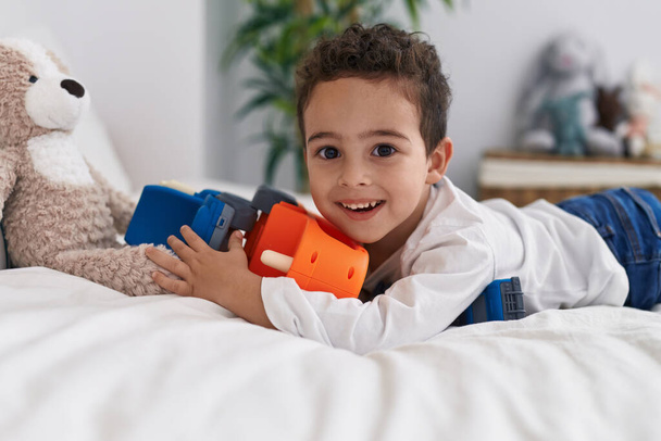 Liebenswerter hispanischer Junge spielt mit Autospielzeug, das im Schlafzimmer auf dem Bett liegt - Foto, Bild