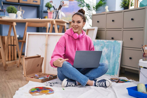 Junges hispanisches Mädchen mit Laptop im Maleratelier lächelt glücklich und zeigt mit Hand und Finger  - Foto, Bild