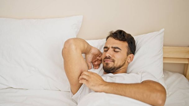 Νεαρός Ισπανός ξαπλωμένος στο κρεβάτι ξύνοντας το χέρι του στην κρεβατοκάμαρα - Φωτογραφία, εικόνα
