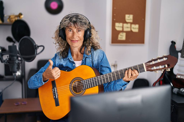 Женщина средних лет играет на классической гитаре в музыкальной студии улыбаясь счастливой и позитивной, большой палец вверх делает отлично и знак одобрения  - Фото, изображение