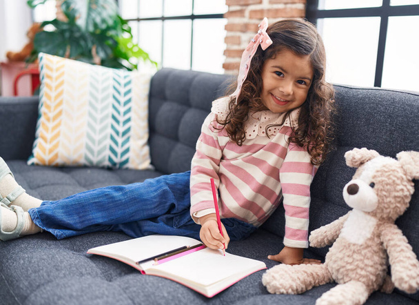 Adorable hispanic girl drawing on notebook sitting on sofa at home - Foto, afbeelding