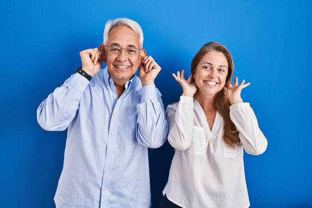 Латиноамериканская пара средних лет, стоящая на синем фоне, улыбаясь, дергая за уши пальцами, смешной жест. проблема прослушивания  - Фото, изображение