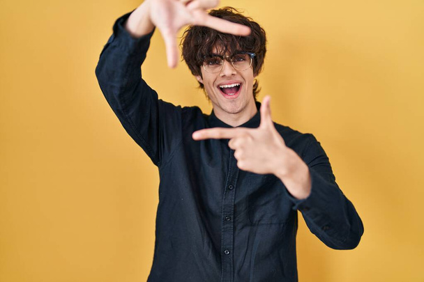 Young man wearing glasses over yellow background smiling making frame with hands and fingers with happy face. creativity and photography concept.  - Zdjęcie, obraz