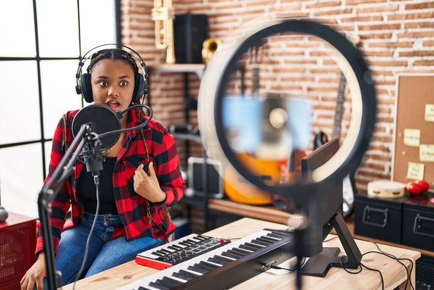 Donna afroamericana con trecce registrazione tutorial musicale con smartphone a casa spaventata e stupita a bocca aperta per sorpresa, faccia incredulo  - Foto, immagini