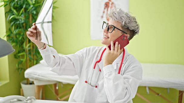 Νεαρή γυναίκα γιατρός μιλάει στο smartphone κοιτάζοντας ακτινογραφία στην κλινική - Φωτογραφία, εικόνα