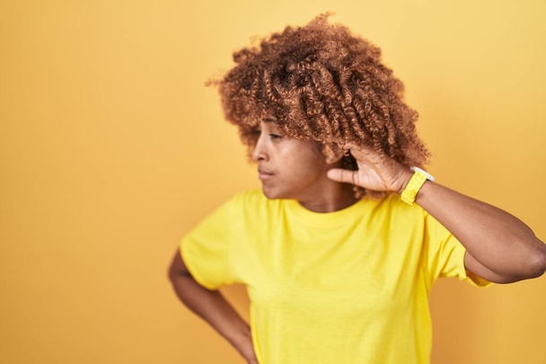 Junge hispanische Frau mit lockigem Haar, die vor gelbem Hintergrund steht und lächelnd mit der Hand über dem Ohr Gerüchten oder Klatsch lauscht. Konzept für Gehörlosigkeit.  - Foto, Bild