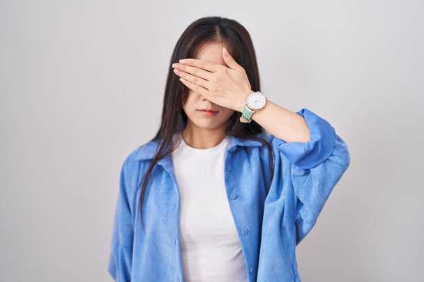 Giovane donna cinese in piedi su sfondo bianco che copre gli occhi con la mano, guardando serio e triste. concetto di avvistamento, nascondiglio e rifiuto  - Foto, immagini