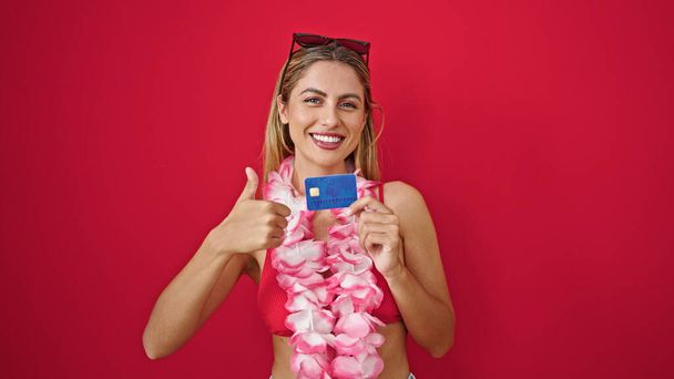 Młoda blondynka turysta trzyma kartę kredytową robi gest w górę nad odizolowanym czerwonym tle - Zdjęcie, obraz