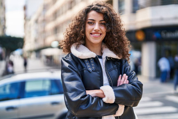Jonge Spaanse vrouw glimlachend vol vertrouwen staand met armen gekruist gebaar op straat - Foto, afbeelding